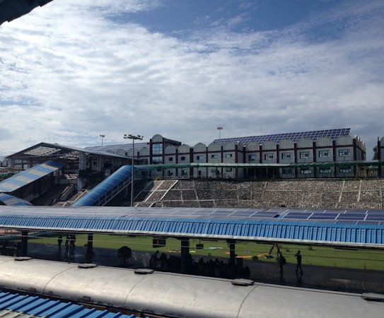 A large structure with numerous solar panels on the roof is visible, suggesting an eco-friendly or energy-efficient building. The building has a modern design with multiple windows and a blue color scheme. A blue, sloped walkway or canopy is present, leading from one section to another. The surrounding area includes green grass and some trees, with a bright, partially cloudy sky overhead.