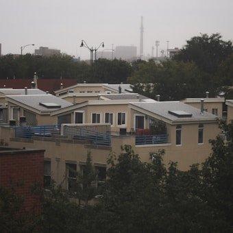 A residential area featuring modern, flat-roofed houses with large windows and solar panels. The homes are surrounded by trees and partially obscured by foliage. In the background, an urban skyline is visible under an overcast sky.