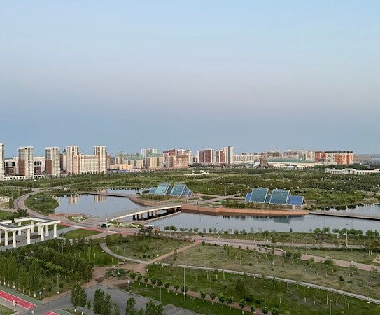 A serene urban landscape featuring high-rise buildings in the background, surrounded by lush greenery and calm water bodies. Solar panels are visible on the banks, indicating a focus on eco-friendly solutions. The area includes paved walkways and well-maintained roads, enhancing accessibility amidst a blend of natural and urban elements.
